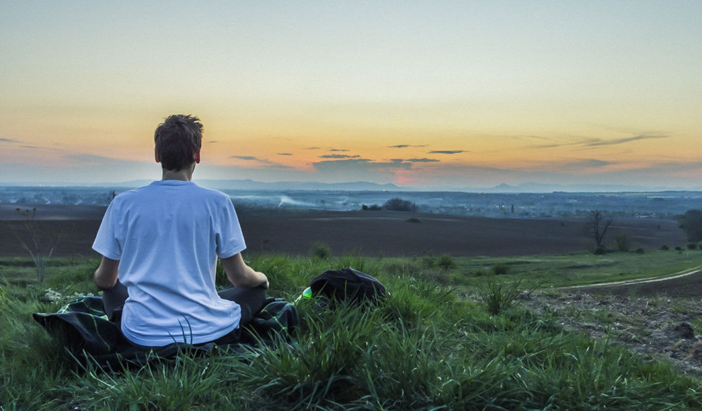 Photo of a person sitting on a grassy landscape, facing the sunset and meditating
